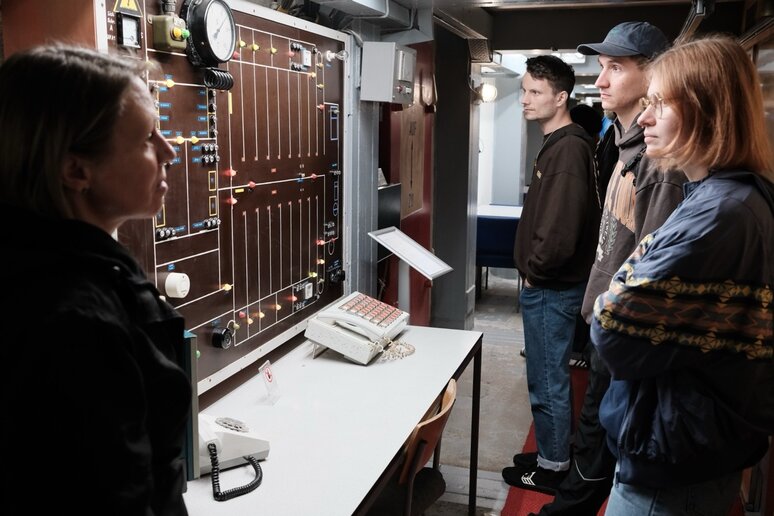 In einem Ausstellungsraum steht links eine Person hinter einem Tisch mit historischer Telefon- und Vermittlungstechnik und zeigt ein Gerät; rechts betrachtet eine kleine Gruppe von Besuchenden die Schaltfelder, auf dem Tisch liegt ein altes Telefon.