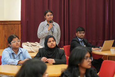 Indische Studierende sitzen an Tischen. Die Studentin in der Mitte steht und spricht in ein Mikrofon.