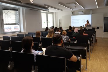 Besetzter Hörsaal mit Teilnehmenden, die einer Kurzvorlesung von Prof.in Wödy zuhören.