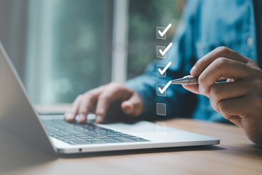 Im Vordergrund ist virtuell eine Checkliste abgebildet. Im Hintergrund sitzt eine Person an einem Tisch mit ihrem Laptop. In der linken Hand hat sie einen Kugelschreiber, mit dem sie Häckchen in die virtuelle Checkliste setzt.