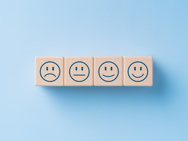 Hellblauer Hintergrund. Darauf in der Mitte vier hölzerne Bauklötze mit Smileys: von schlecht gelaunt zu gut gelaunt.