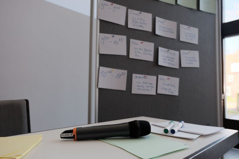 Auf einem Seminartisch liegen ein Handmikrofon, Moderationskarten und zwei Stifte. Im Hinterrund ist ei Whiteboard mit beschrifteten Karten. Auf diesen stehen Uhrzeiten, Themen und Namen.