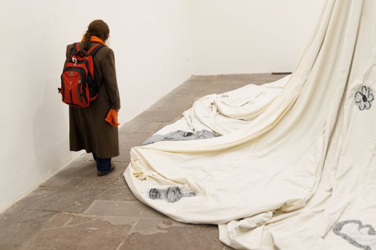 Eine Frau von hinten. Sie schaut sich ein Ausstellungsstück an. Ein großes weißes Tuch mit schwarz-weißen Aufnähern von Kindern.