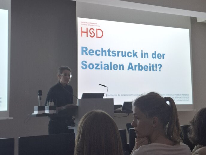 Ein Frau steht an einem Redepult. Hinter ihr ist der Titel des Vortrags "Rechtsruck in der Sozikalen Arbeit?!" und das Logo der Hochschule Düsseldorf projiziert.