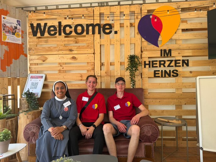 Drei Personen sitzen auf einer Couch, eine trägt Nonnentracht und die anderen beiden ein rotes Poloshirt. Auf der Holzwand hinter ihnen steht: Welcome. Im Herzen eins.