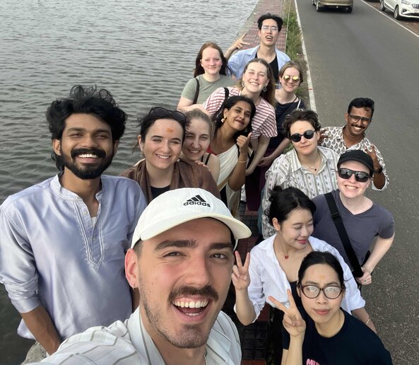 Selfie einer Gruppe von 14 Erwachsenen auf einer Uferpromenade; alle lächeln in die Kamera, links ist Wasser sichtbar.