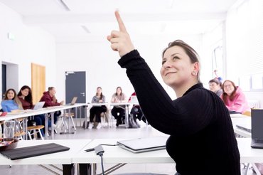 Eine Studentin im Seminarraum zeigt mit ihrem Finger auf etwas oberhalb des Bildausschnittes. Im Hintergrund sieht man weitere Studierende an Tischen, die zur Studentin schauen.