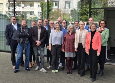 Gruppenfoto von etwa 15 Personen unterschiedlichen Alters und Geschlechts in Business‑Casual, lächelnd und in zwei Reihen vor der Glasfassade eines Hochschulgebäudes.