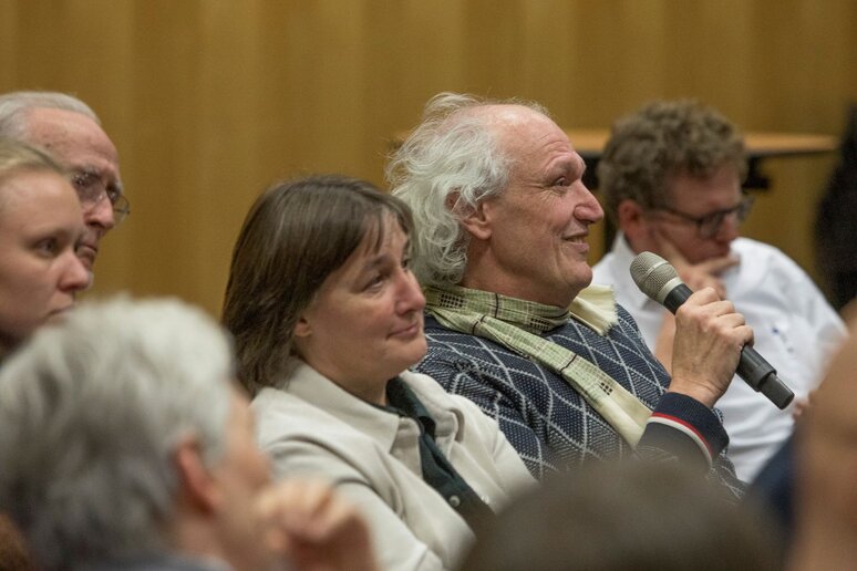 Blick in eine Stuhlreihe in der Menschen sitzen, die letzte Person hat ein Mikrofon in der Hand.