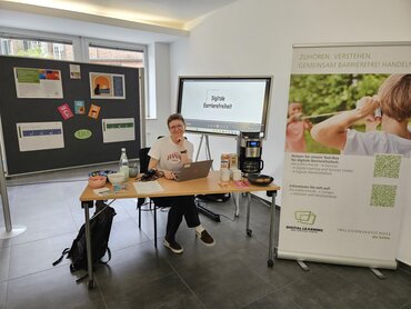 Das Bild zeigt eine Frau an einem Tisch sitzend auf dem Informationsmaterial liegt. Im Hintergrund ist ein Roll up mit Infos zum GAAD.
