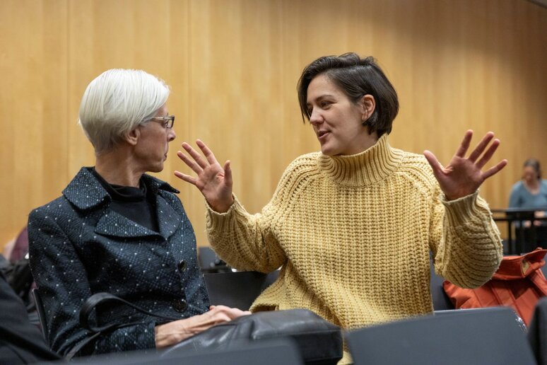 Zwei Frauen unterhalten sich. Die junge Frau im gelben Pullover erklärt der anderen Frau etwas und gestikuliert dabei mit ihren Händen in der Luft.