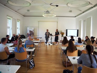 Teilnehmende sitzen an Tischreihen im Seminarraum. Vor Kopf stehen Professor Laukötter und Professorin Panreck.