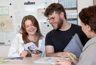 Eine Studentin und ein Student stehen an einem Tisch und schauen gu gelaunt in einen katho-Flyer hinein. Vor ihnen am Rand steht die Studienberaterin.