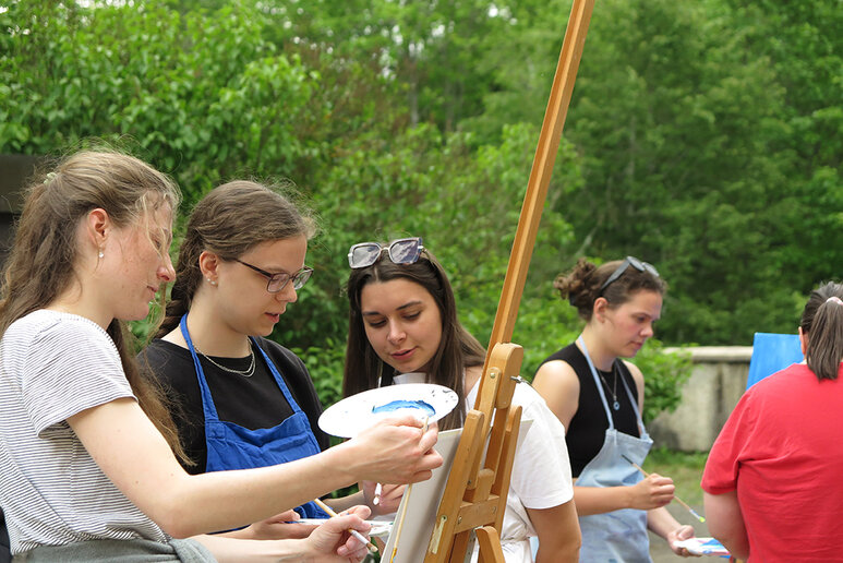 Drei Frauen malen gemeinsam an einem Bild, welches auf einem Stativ steht. Das Bild selbst ist nicht zu sehen.