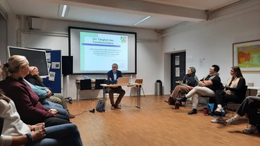 Am Kopfende sitzt ein Mann und referiert, um ihn herum ist ein Stuhlkreis mit Menschen