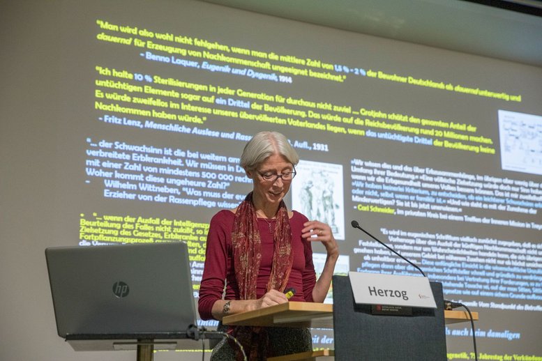 Eine Frau steht hinter einen Rednerpult. Im Hintergrund sieht man eine Powerpointfolie auf der mehrere Zitate abgebildet sind.