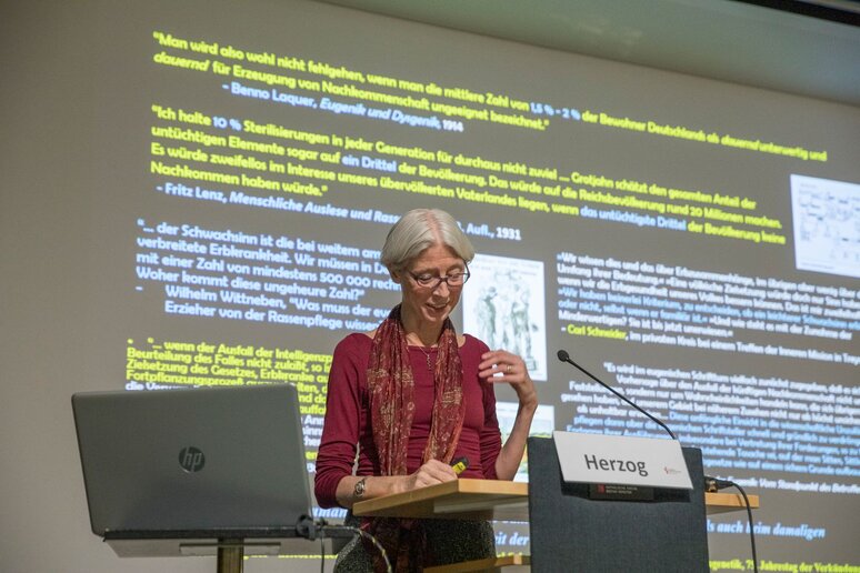 Eine Frau steht hinter einen Rednerpult. Im Hintergrund sieht man eine Powerpointfolie auf der mehrere Zitate abgebildet sind.