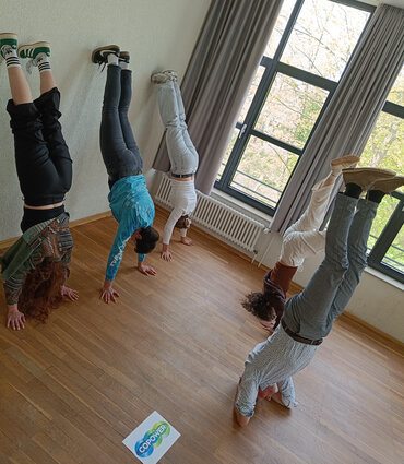 Fünf Personen machen im Rahmen der CoPoweR-Methode einen Handstand mit Wandkontakt in einem hellen Seminarraum, während im Vordergrund das CoPoweR-Logo auf dem Boden liegt. Die Übung vermittelt spielerisch Vertrauen und Teamgeist für traumasensible Konfliktarbeit.