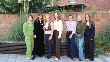 Gruppenfoto von sieben Erwachsenen im Freien vor einer Backsteinmauer mit hohen Ziergräsern; alle blicken lächelnd in die Kamera.