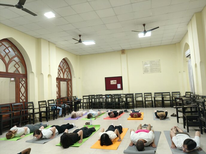 Mehrere Personen liegen entspannt auf Yogamatten in einem großen, hellen Saal; Stühle sind an den Wänden aufgestellt.