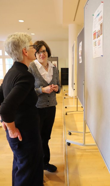 Zwei Frauen stehen vor einer Reihe Pinnwänden mit Plakaten. Eine der beiden schaut auf das Plakat und spricht, die andere hört ihr zu.
