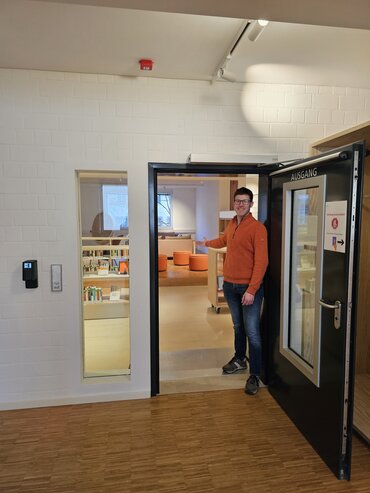 Blick durch die geöffnete Bibliothekstür in die Bibliothek. Ein Mitarbeiter steht in der Tür und weist mit freundlicher Geste in die Bibliothek.