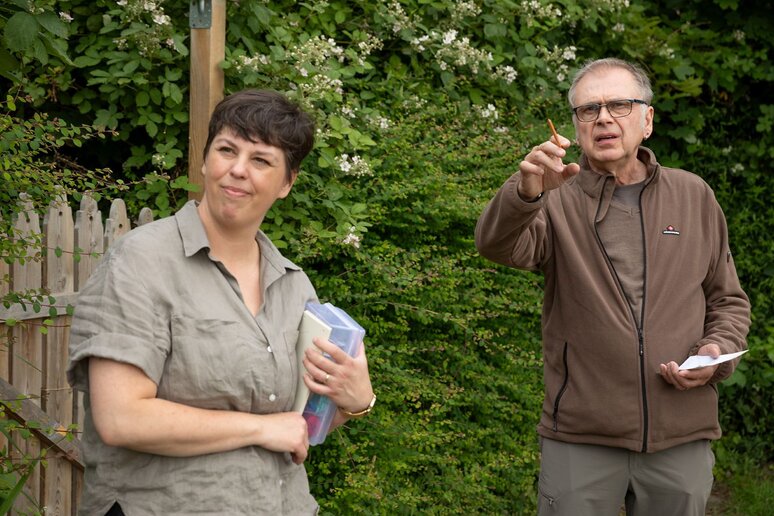 Zwei Personen ssitzen nebeneinander, die eine Person zeigt in Richtung der Kamera.