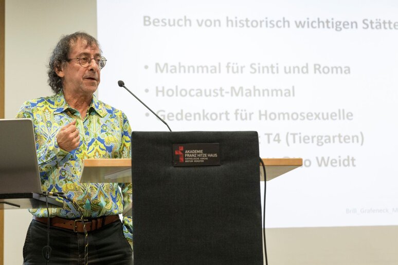 Ein Mann in sehr buntem Hemd steht am Rednerpult. Im Hintergrund eine Präsentation über verschiedene Mahnmale von Opfern des Holocaust.