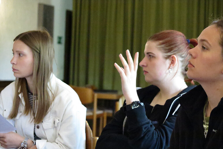 Drei Frauen sitzen nebeneinander und schauen in die gleiche Richtung. Die Frau in der Mitte hebt ihre Hand für eine Wortmeldung.