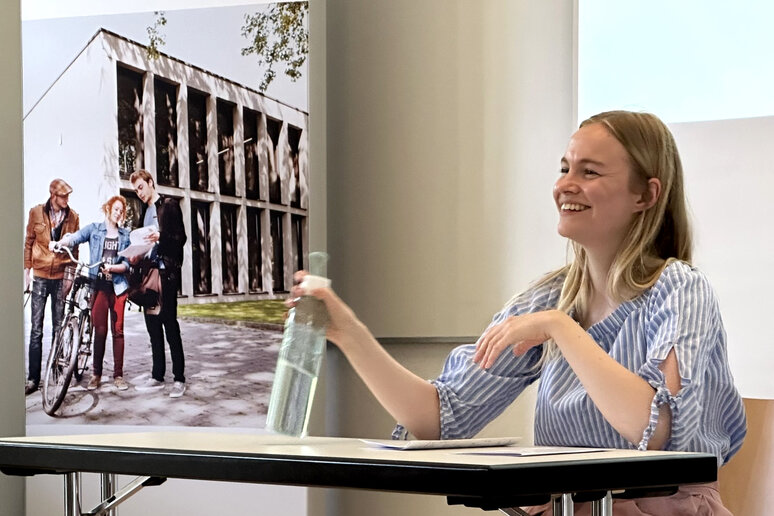 Sarah Althöfer sitzt an einem Tisch und lächelt. Neben ihr steht ein katho-Roll-up.
