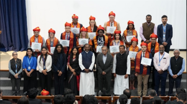 Gruppenfoto auf einer Bühne bei einer Zertifikatsverleihung. Etwa 25 Personen stehen in zwei Reihen; in der hinteren Reihe tragen mehrere Personen orangefarbene Schärpen und rote Kopfbedeckungen und halten Urkunden.