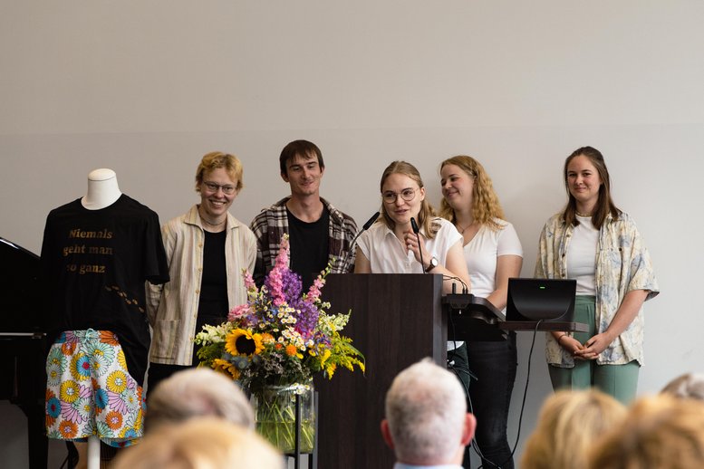 Eine Gruppe von Studierenden steht auf der Bühne; eine von ihnen hält am Pult eine Rede. Das Geschenk - ein von Studierenden unterzeichnetes T-Shirt - wird auf einer Schaufensterpuppe präsentiert.