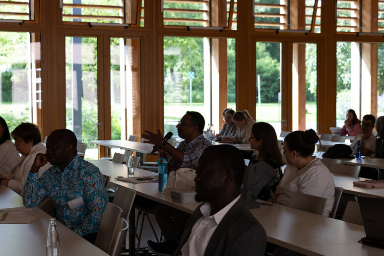 Eine Person sitzt im Vorlesungssaal und hat ein Mikrofon in der Hand. Die Person spricht zu allen anderen Zuhörenden. 