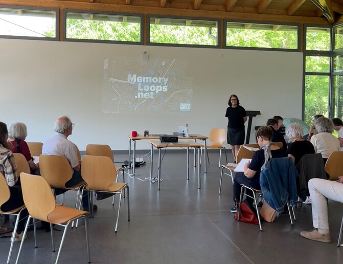 Eine Frau steht vor einer Leinwand auf der "Memory Loops.net" steht. Ein Gruppe Menschen sitzt davor.