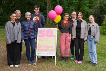 Acht Menschen stehen auf einer Wiese, eine Person hat drei Luftballons in der Hand. Vor ihnen steht ein Werbeplakat für die Mülheimer Theatertage.