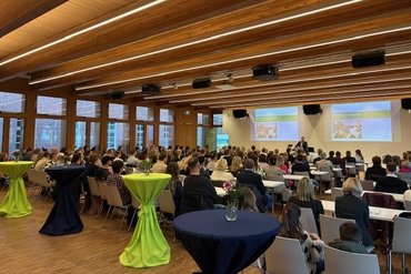 Ein voller Saal ist zu sehen. Eine Person steht vorne und hält eine Begrüßungsrede, viele Personen hören zu