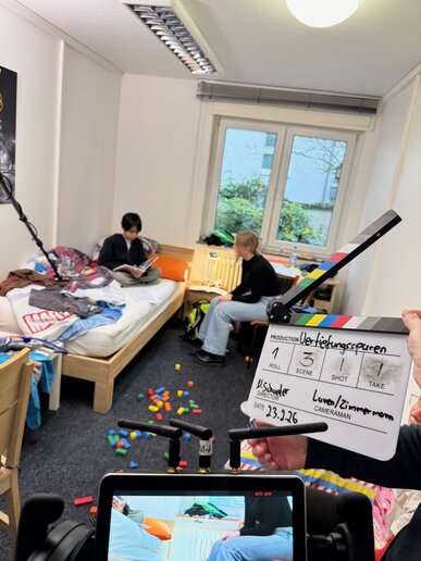 Eine Person sitzt lesend auf einem Bett; eine zweite Person sitzt auf einem Stuhl vor dem Bett. Auf dem Boden liegen Bauklötze