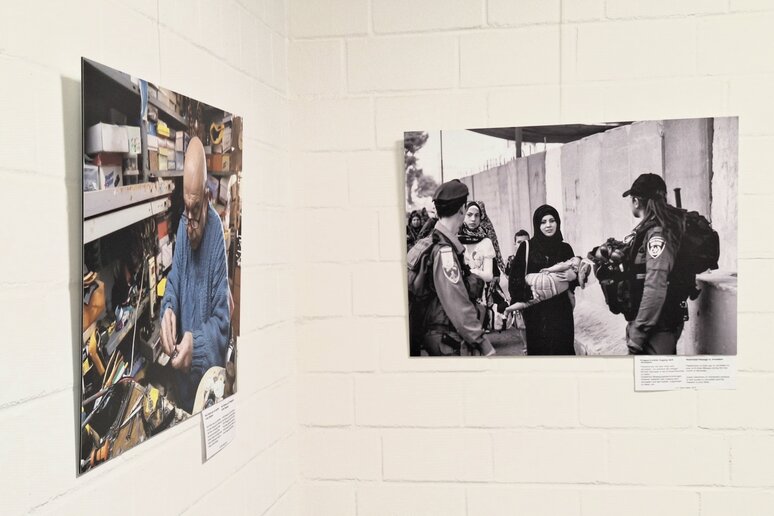 Zwei großformatige Bilder hängen über Eck an einer Wand. Links sitzt ein Mann in seiner Werkstatt, rechts treffen Frauen und ihre Kinder an einer Grenzmauer auf zwei Soldaten.