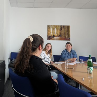 Drei Personen sitzen an einem Tisch in einem Raum der katho und unterhalten sich. 