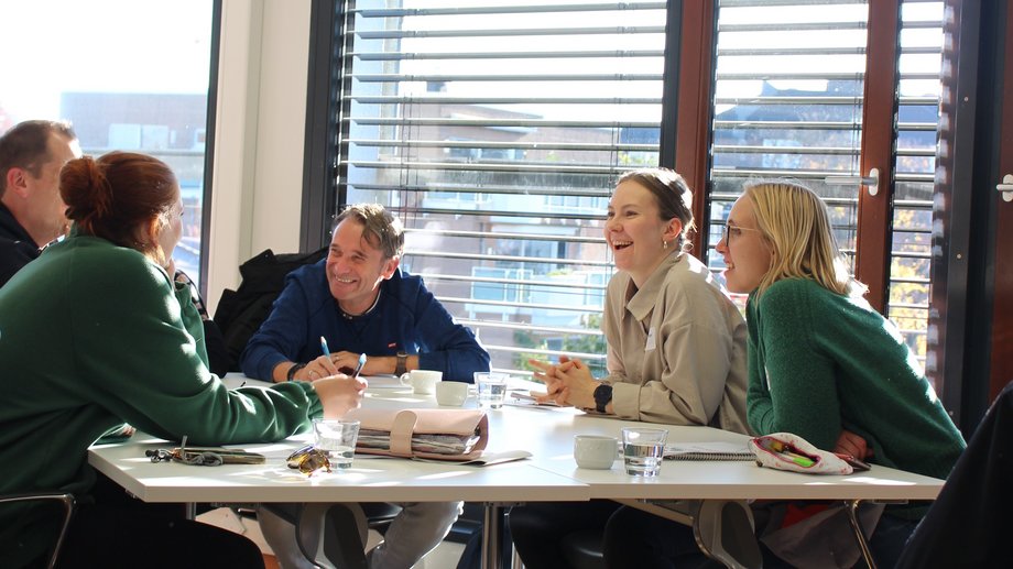 Sechs Erwachsene sitzen in einem hellen Seminarraum um einen Tisch, sprechen und lachen; auf dem Tisch liegen Notizbücher und Tassen, Sonnenlicht fällt durch Jalousien.