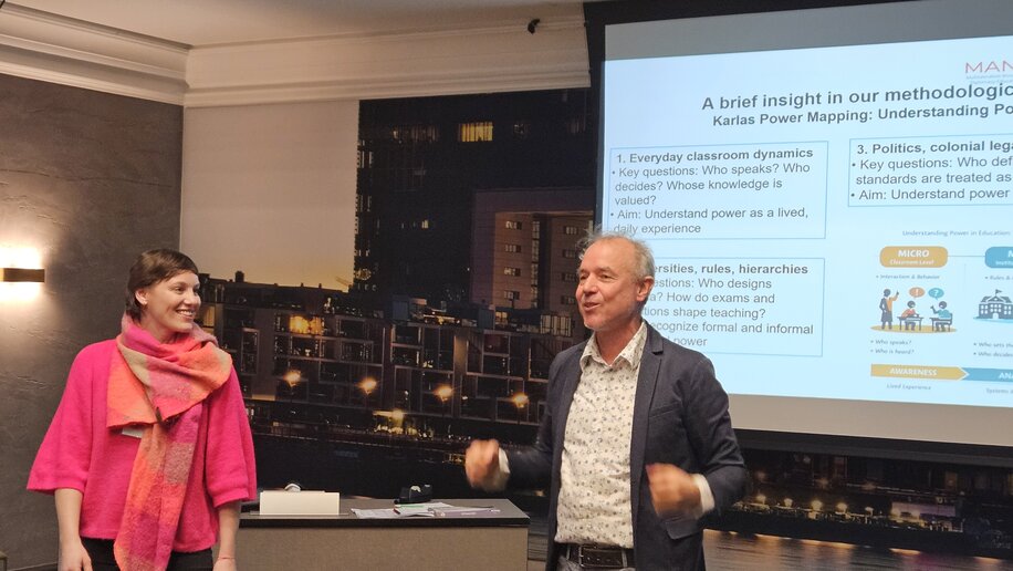 Zwei Personen stehen vor einer Präsentationsleinwand in einem Raum. Die Frau trägt einen pinkfarbenen Schal und lächelt, während der Mann gestikulierend spricht. Auf der Leinwand ist eine Präsentation mit dem Titel „A brief insight in our methodologies“ zu sehen, die Themen wie „Everyday classroom dynamics“ und „Politics, colonial legacies“ behandelt. Der Raum ist professionell eingerichtet und die Atmosphäre scheint locker und informativ zu sein.
