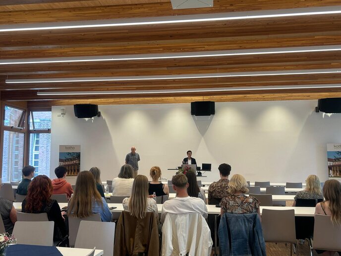Prof. Dr. Sabine Engel und Prof. Dr. Klaus Hartmann moderieren auf der Bühne. Die Zuschauer hören zu.