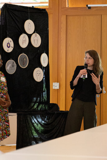 Eine Frau steht vor einem Kunstobjekt, sie hat ein Mikrofon in der Hand und stellt das Kunstwerk vor.  ein KUnstprojekt vor.