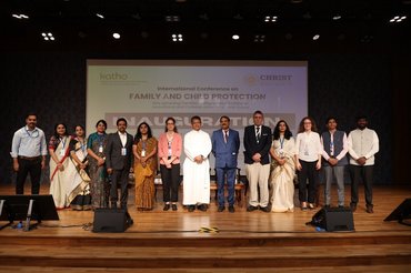 Eine Gruppe von Menschen steht auf einer Bühne vor einer Leinwand, die eine internationale Konferenz über Familien- und Kinderschutz ankündigt. Sie sind formell gekleidet, und einige tragen Namensschilder. Die Veranstaltung scheint Teil einer feierlichen Eröffnung zu sein.