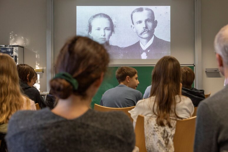 Einige Menschen sitzen in einem Klassenraum und schauen Richtung Tafel. Über der Tafel ist ein schwarz-weiß Bild von einem Mann und einer Frau.