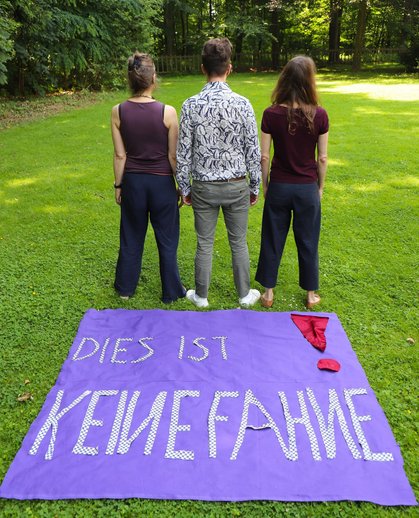 Auf eienr Wiese liegt ein großer lila Stoff mit dem Schriftzug "Dies ist keine Fahne" und einem roten Ausrufezeichen. Dahinter stehen drei Studierende mit dem Rücken zur Fahne, sie wenden sich bewusst ab.