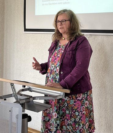 Prof.in Dr.in Silvia Hamacher steht auf der Bühne und hält ihren Vortrag in Form einer PowerPoint Präsentation, die an der Wand hinter ihr zu sehen ist.