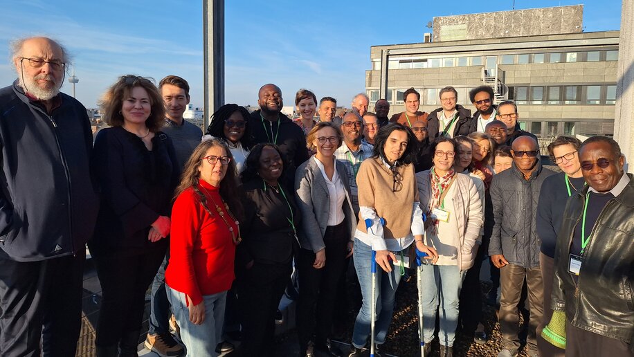 Eine Gruppe von Menschen posiert draußen für ein Foto auf einer Dachterrasse bei schönem Wetter. Sie lächeln in die Kamera, einige tragen Lanyards. Im Hintergrund ist ein Gebäude zu sehen. Die Atmosphäre wirkt freundlich und der Himmel ist klar und blau.