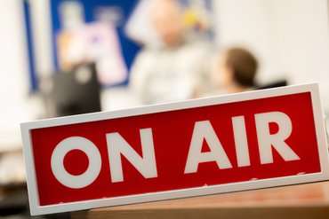 Ein rotes Schild mit Aufschrift On Air steht im Vordergrund. Im Hintergrund stehen verschwommen zwei Personen.