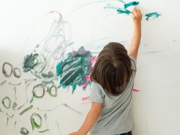 Ein Kind steht vor einer Wand und bemalt diese mit einem Pinsel. Zu sehen sind dunkelgrüne Kreise und schraffierte Flächen.